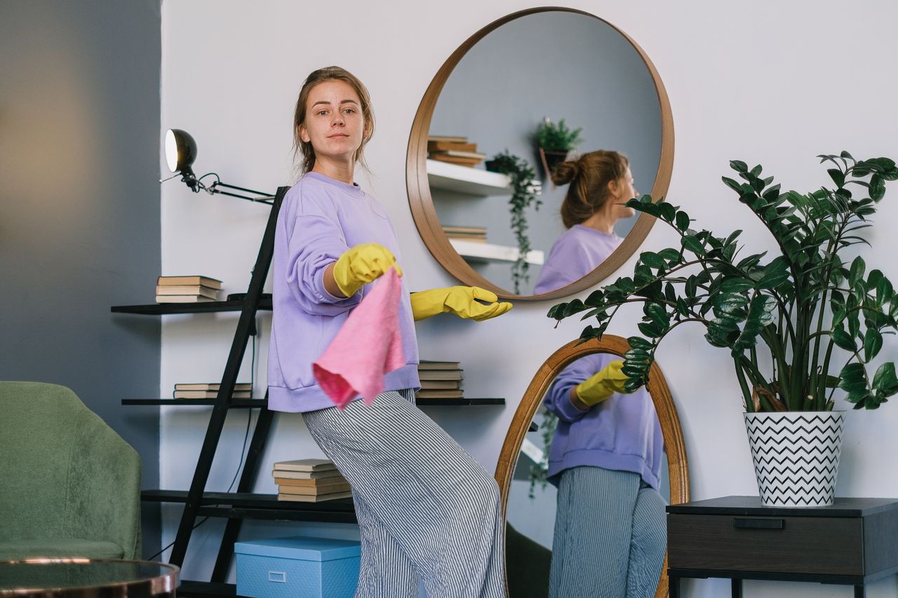 Side view of cheerful woman in casual clothes and latex gloves standing near mirrors while cleaning apartment in daytime